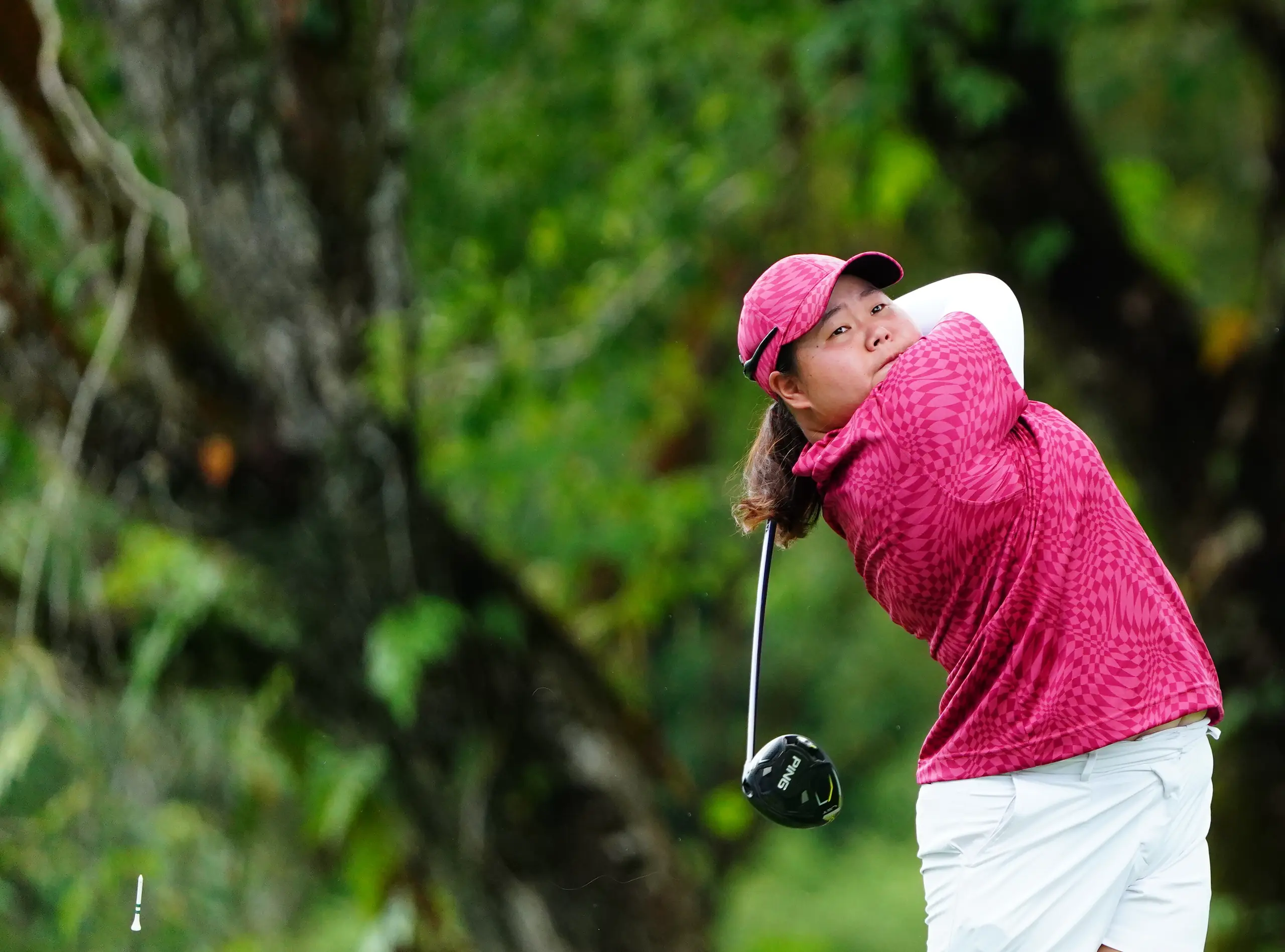A golfer follows through on her tee shot, framed by tall trees
